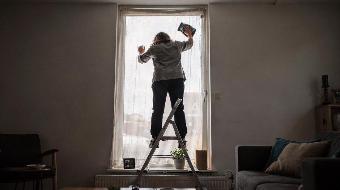 Person on a small folding step stool reaching the top of a tall window frame in a Dutch living room