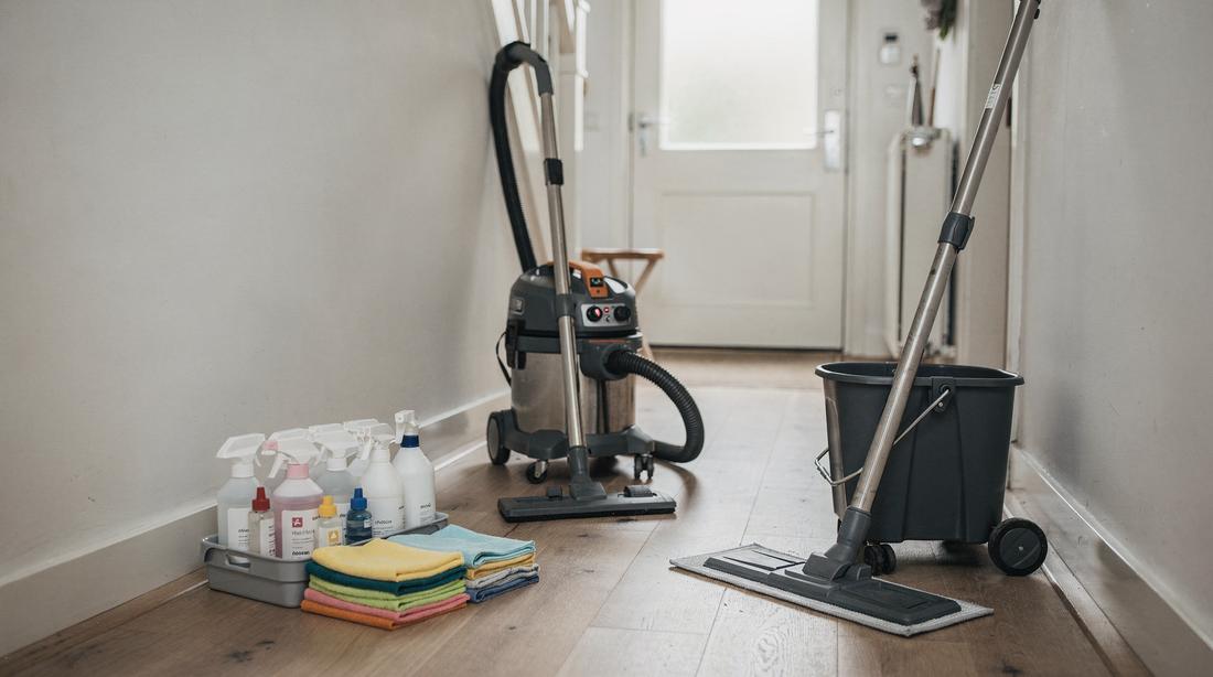 Professional cleaning equipment in a Dutch hallway: commercial vacuum, caddy of labelled bottles, stacked colour-coded cloths
