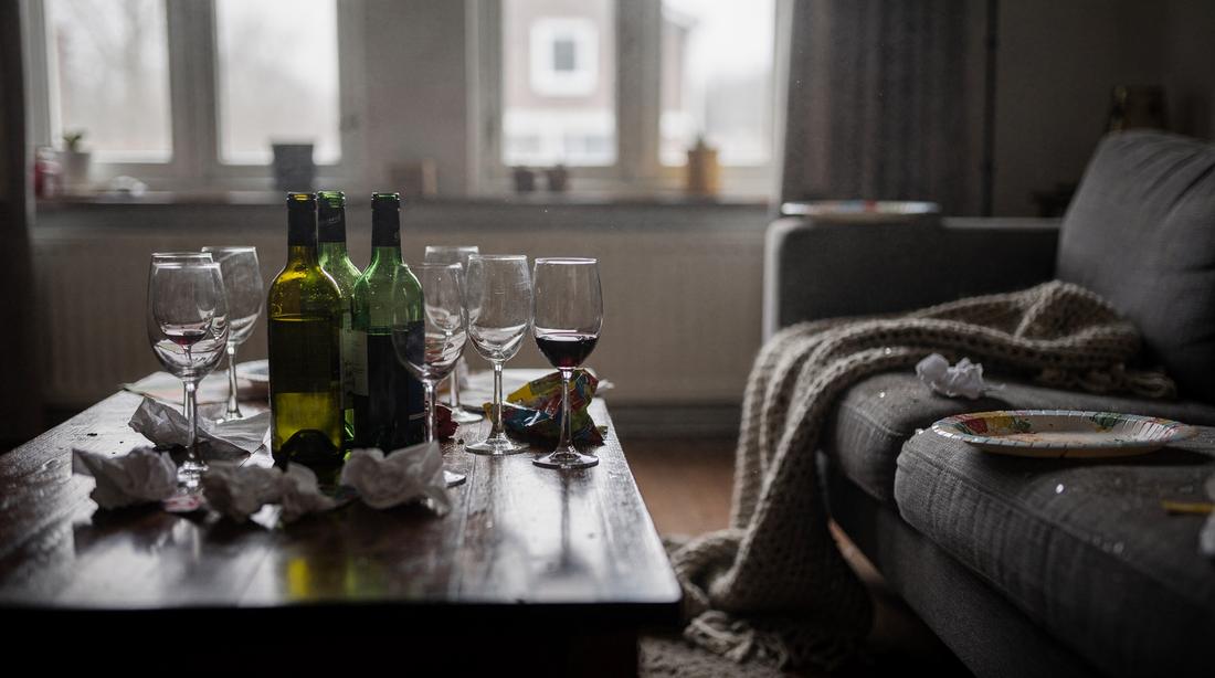 Woonkamer de ochtend na een feest: wijnglazen en flessen op een houten tafel, servetten en een plaid waar ze gevallen zijn